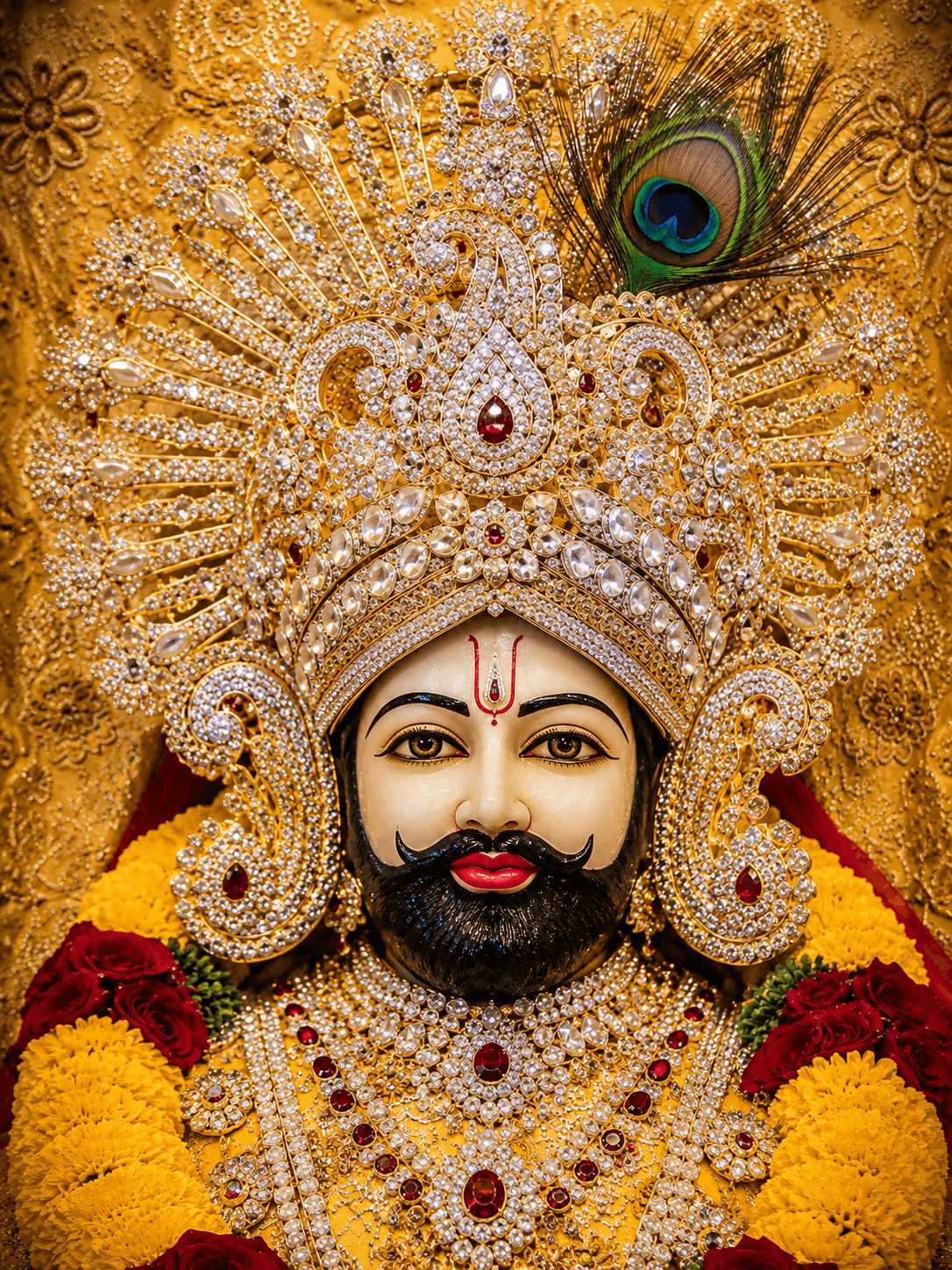 Beautiful 4K image of Khatu Shyam Ji with a grand diamond-studded crown, peacock feather, ruby jewelry, and floral garlands, free HD wallpaper download.